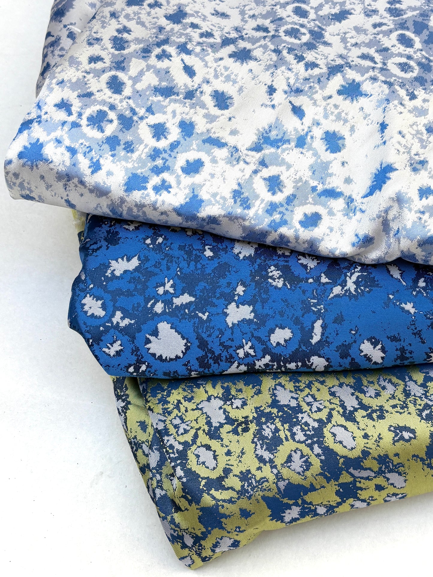 Stack of three folded silk samples with blue, white, and yellow floral patterns on a white background.