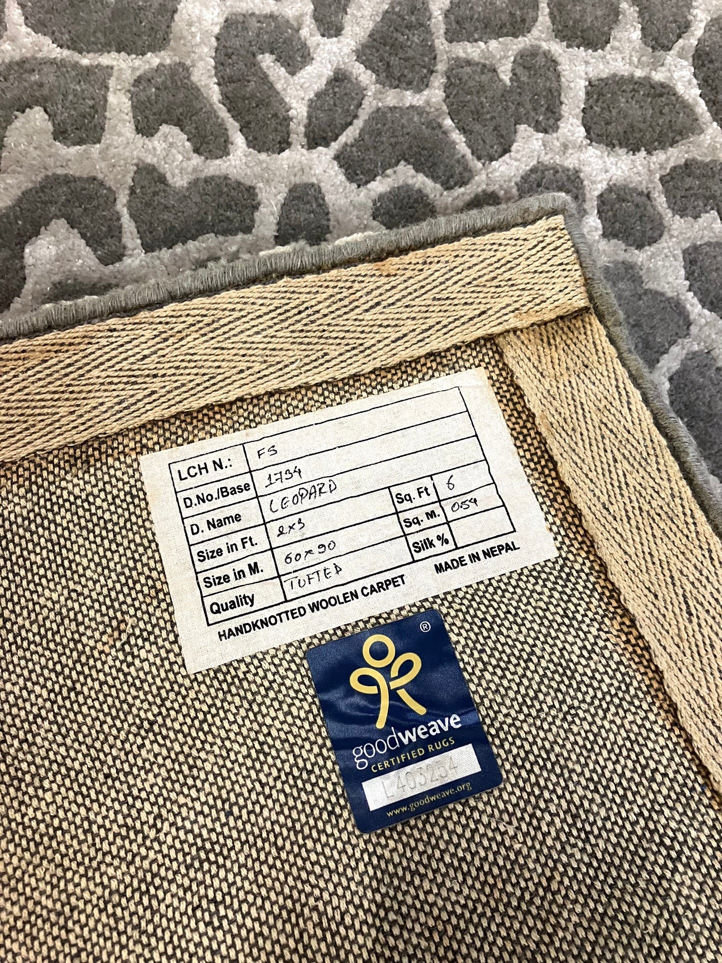Close-up of the back of a rectangular, hand knotted wool rug with a leopard animal print pattern in shades of brown and beige, with a sticker that contains the content of its.