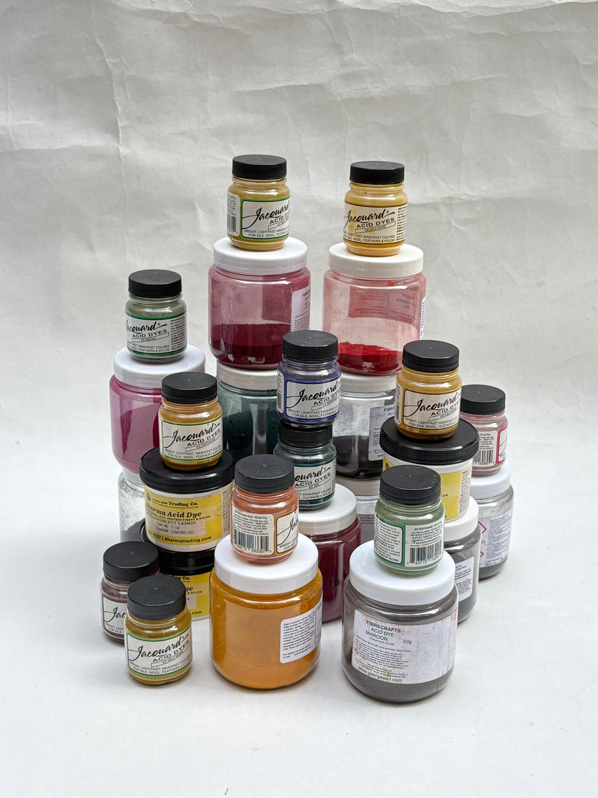 A collection of small jars of yarn dye pigments paint in various colours, arranged in a pyramid-like stack on a white background.