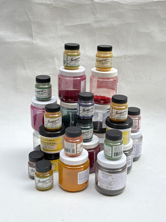 A collection of small jars of yarn dye pigments paint in various colours, arranged in a pyramid-like stack on a white background.