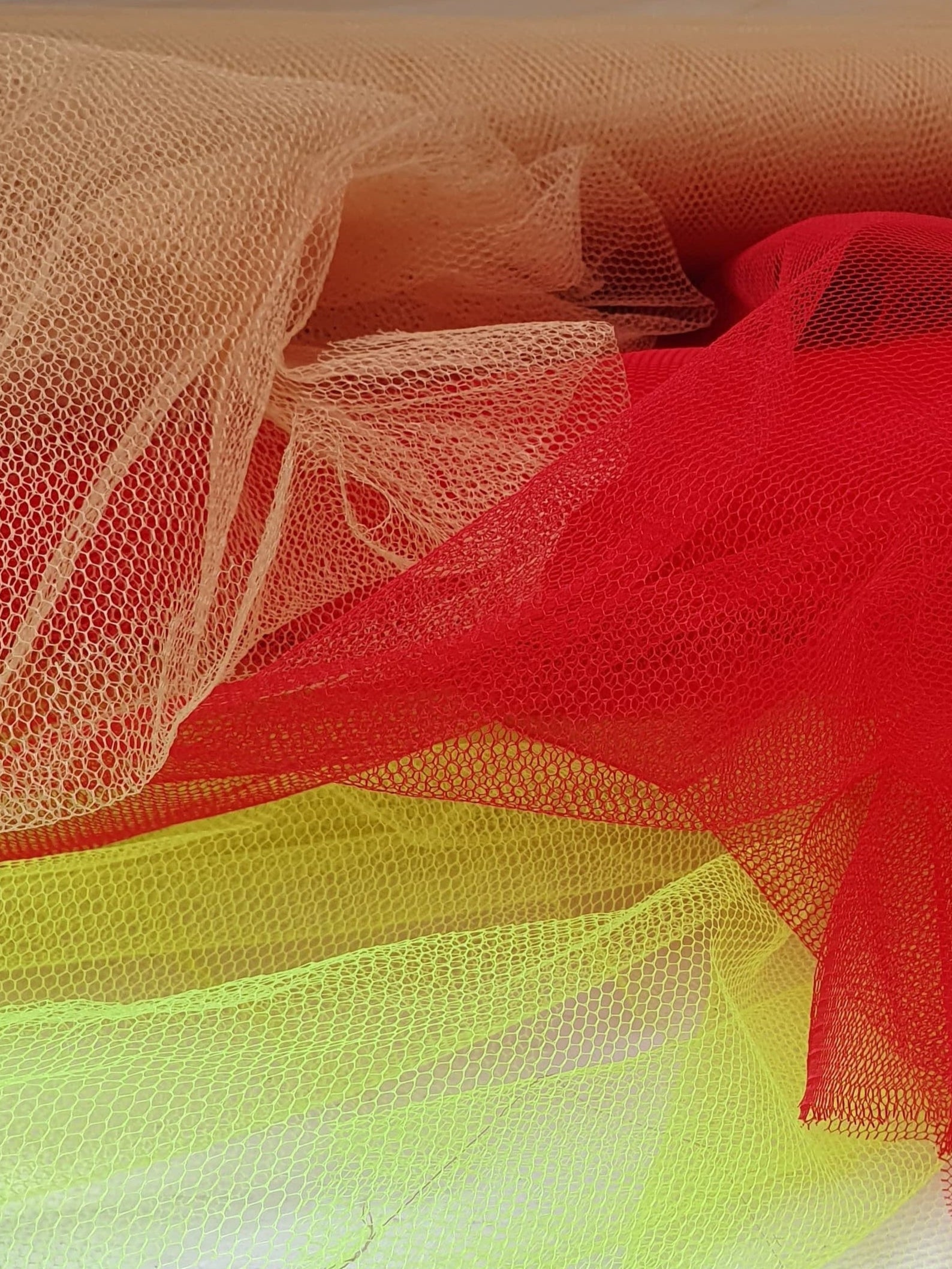 A close-up of voile tulle fabric in neon yellow, cinnamon, and red, showcasing its breathable net texture.