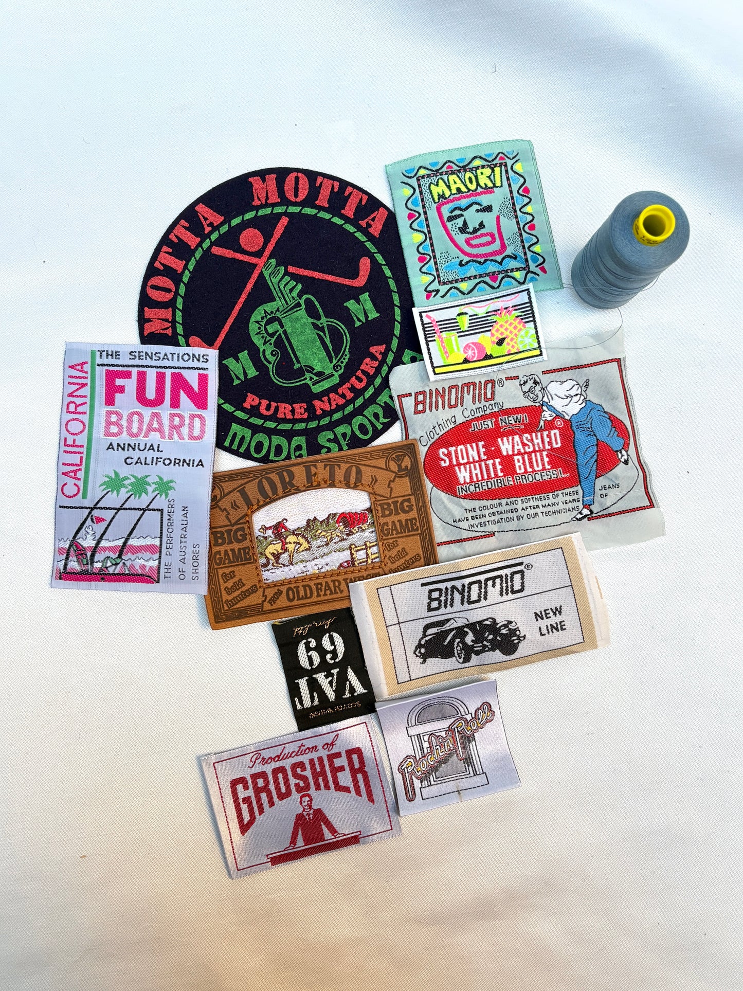Assorted woven badges and labels arranged beside a grey thread spool.