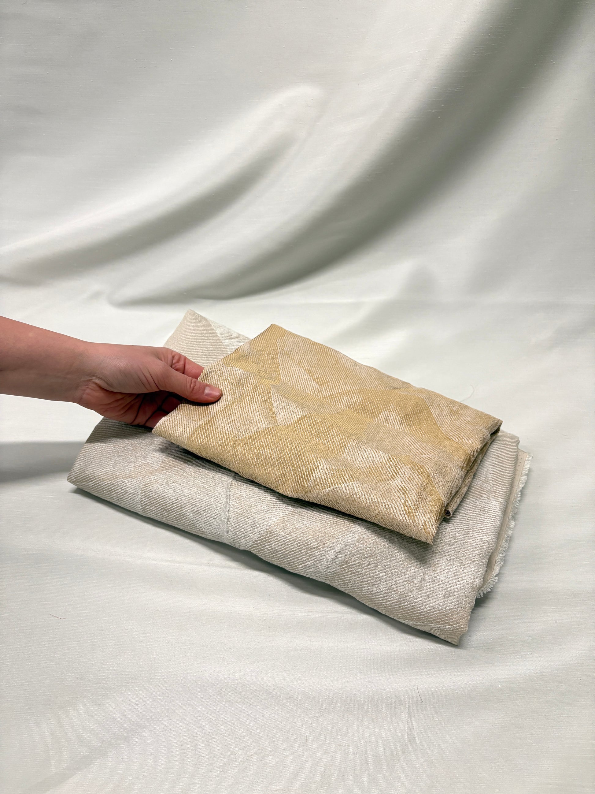 Hand holding a folded beige fabric over another fabric on a white background