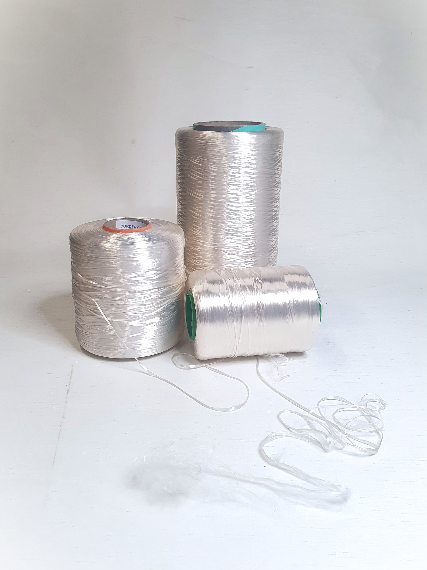 Three cones of rayon multifilament yarn with different thicknesses placed on a light background, along with a small piece of the yarn showing its texture.